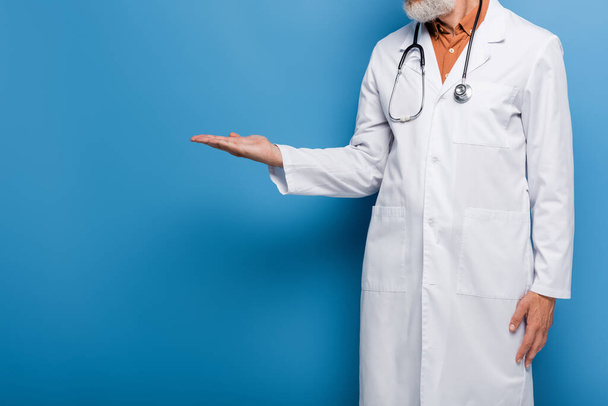 vista recortada del médico barbudo de mediana edad en bata blanca apuntando con la mano sobre azul - Foto, imagen