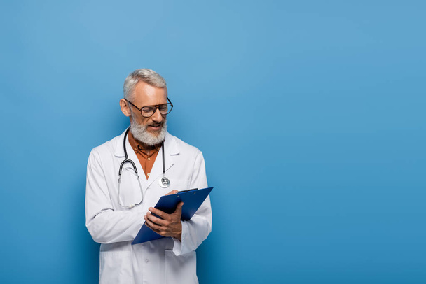 médico de mediana edad en abrigo blanco y gafas de escribir prescripción en portapapeles en azul - Foto, Imagen