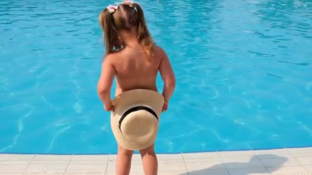 klein meisje staat en bedekt zich met een zonnehoed tegen de achtergrond van het blauwe water van het zwembad of de zee. Het concept van zomervakantie, zonnebrandcrème, reizen. selectieve aandacht voor hoed - Video
