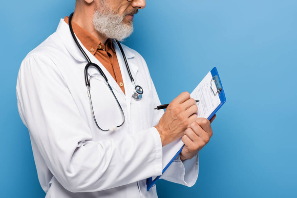 cropped view of middle aged doctor in white coat writing prescription on clipboard on blue - Foto, Imagen