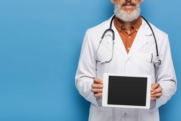cropped view of bearded middle aged doctor in white coat holding digital tablet with blank screen on blue  - Zdjęcie, obraz