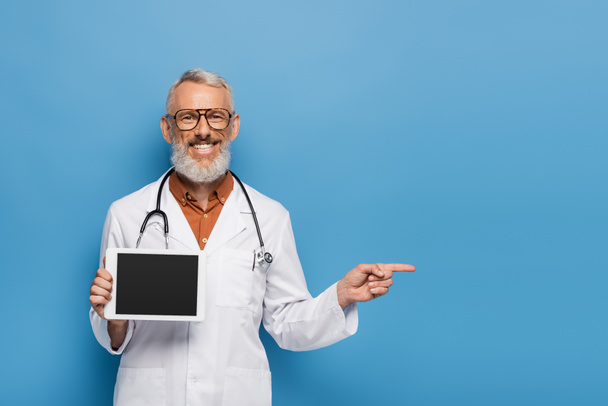 happy middle aged doctor in white coat holding digital tablet with blank screen while pointing with finger on blue  - Фото, зображення