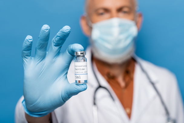 wazig arts in medische masker met flacon met coronavirusvaccin geïsoleerd op blauw - Foto, afbeelding