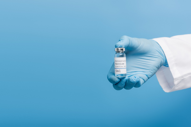 bijgesneden zicht op arts in latex handschoen met flacon met coronavirusvaccin geïsoleerd op blauw - Foto, afbeelding