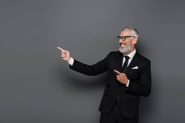 joyful middle aged businessman in suit and glasses pointing with fingers on grey - Fotó, kép