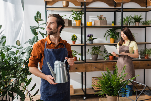 Young florist holding watering can near plants and blurred colleague in flower shop  - Foto, Bild