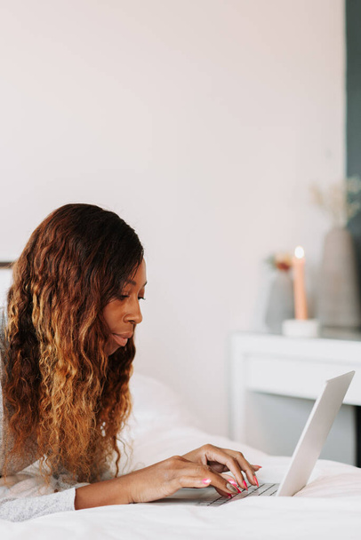 Egy fiatal, gyönyörű fekete nő az ágyon, otthonról dolgozik a laptopján. - Fotó, kép