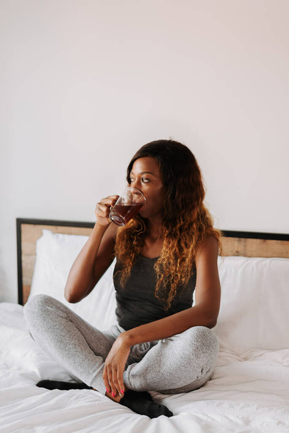 A young beautiful black female relaxing sitting on the bed drinking a cup of coffee - Фото, изображение