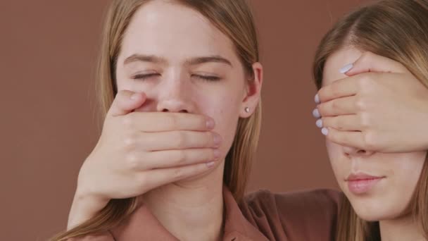 Panning close up ritratto di giovani donne gemelle che coprono la bocca e gli occhi, mentre in posa per la fotocamera contro sfondo marrone - Filmati, video