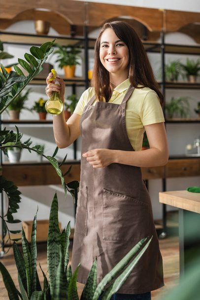 Positive florist holding sprayer near plants and looking at camera in flower shop  - Foto, imagen