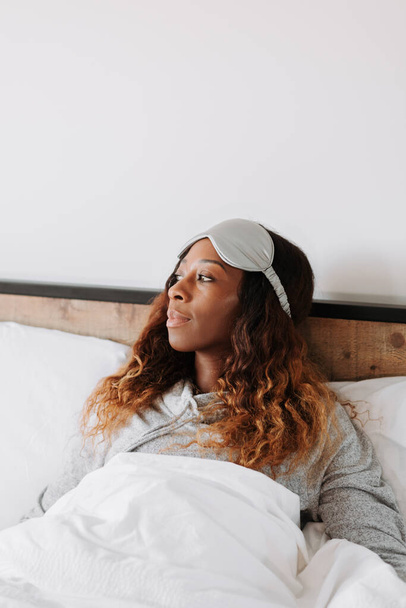A vertical shot of a black female lying under a blanket in pajamas with an eye sleep mask - Foto, Imagen