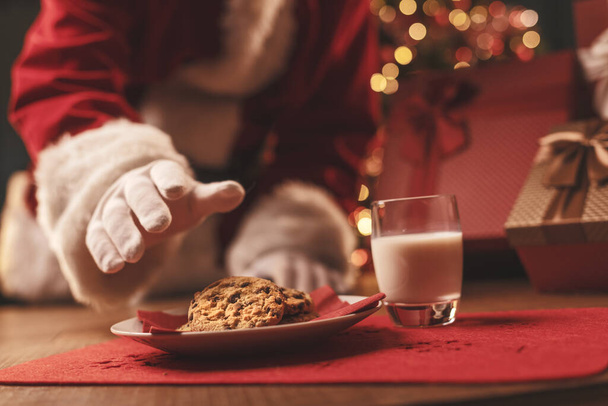 Santa Claus having a delicious snack, he is eating cookies and drinking milk, Christmas and holidays concept - Foto, afbeelding