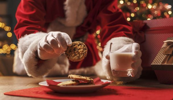 Santa Claus having a delicious snack, he is eating cookies and drinking milk, Christmas and holidays concept - Photo, image