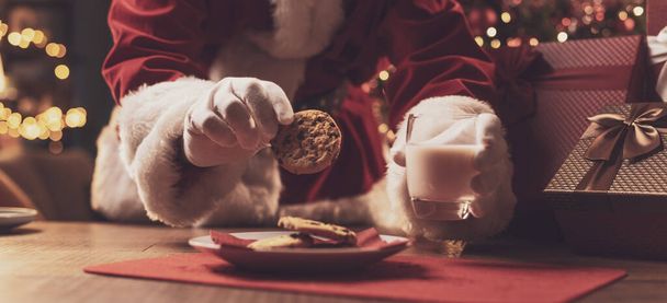 Santa Claus having a delicious snack, he is eating cookies and drinking milk, Christmas and holidays concept - Foto, imagen