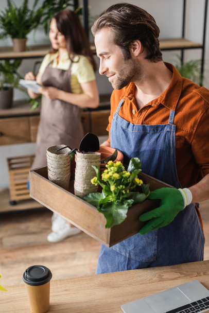 Young florist holding box with gardening tools and flowerpots near laptop and coffee in flower shop  - Photo, Image