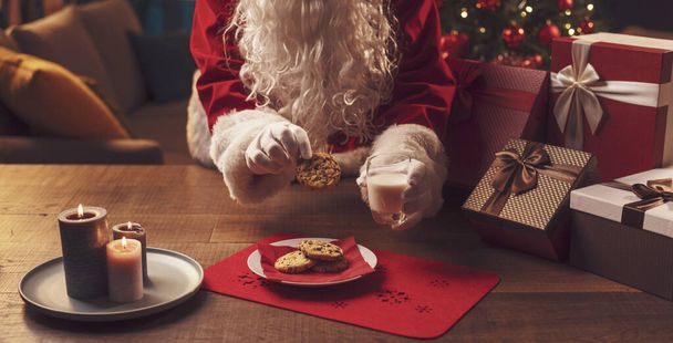 Santa Claus having a delicious snack, he is eating cookies and drinking milk, Christmas and holidays concept - Фото, изображение