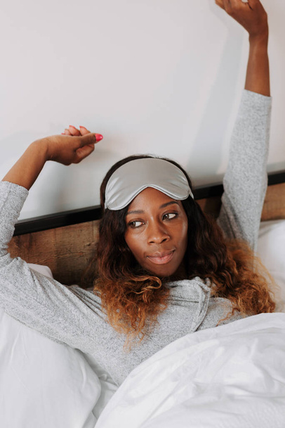 A vertical shot of a black female lying under a blanket in pajamas with an eye sleep mask - Foto, immagini