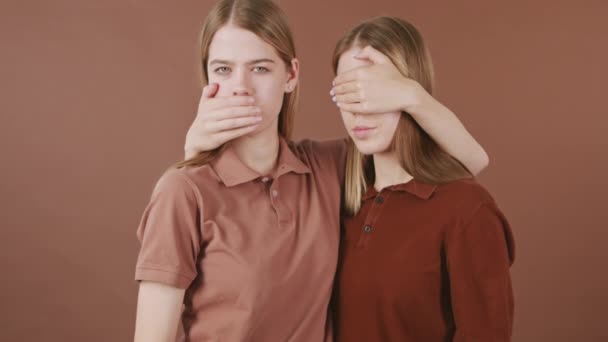 Retrato inclinado de hermanas gemelas idénticas de pie sobre fondo marrón y cubriendo la boca y los ojos - Metraje, vídeo