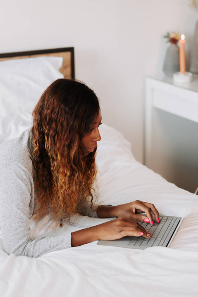 A young beautiful black female on the bed working from home on her laptop - Fotó, kép