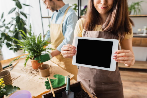 Florist in apron holding digital tablet with blank screen near blurred colleague and plants in flower shop  - Photo, Image