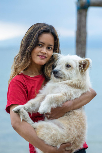A vertical shot of an Indonesian woman hugging a dog on the shore in Togian Island - Photo, image