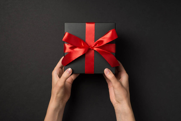 First person top view photo of female hands holding black giftbox with vivid red ribbon bow on isolated black background - Foto, Bild