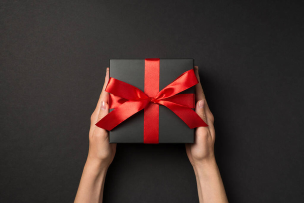 First person top view photo of hands holding stylish black giftbox with red satin ribbon bow on isolated black background - Photo, Image