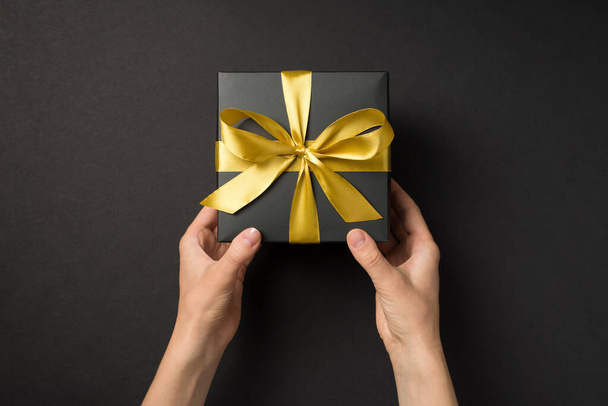 First person top view photo of hands giving black giftbox with golden satin ribbon bow on isolated black background - Фото, зображення
