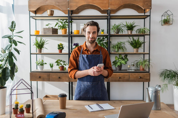 Florist holding smartphone near laptop and coffee in flower shop  - Foto, afbeelding