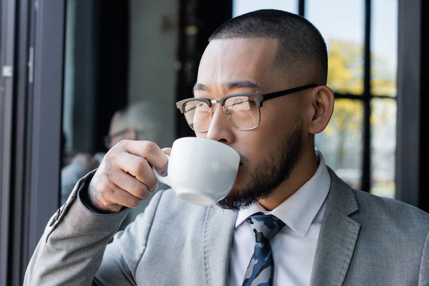 asian businessman in formal wear and eyeglasses drinking coffee in office - Photo, Image
