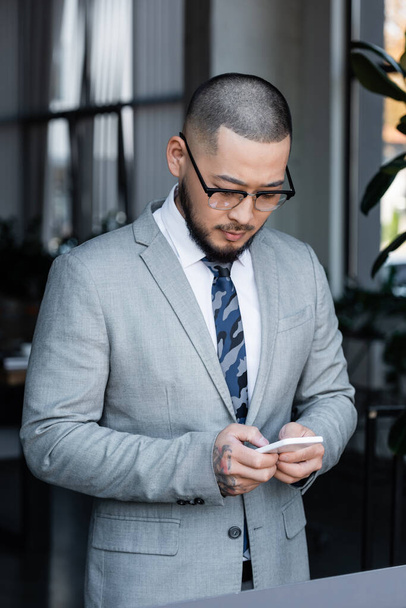 asian businessman in formal wear and eyeglasses messaging on cellphone in office - Foto, Bild