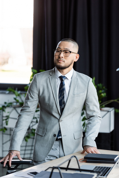 asian businessman in formal wear and eyeglasses looking away while standing at work desk - Fotografie, Obrázek
