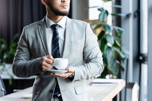 vista ritagliata di uomo d'affari barbuto in abito formale in piedi con una tazza di caffè in ufficio - Foto, immagini