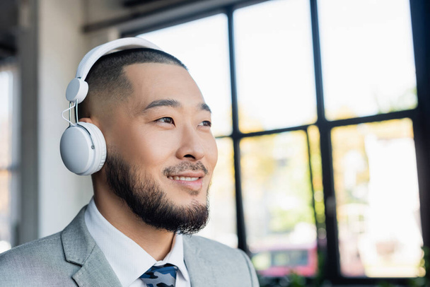 happy asian businessman in wireless headphones looking away in office - Фото, изображение