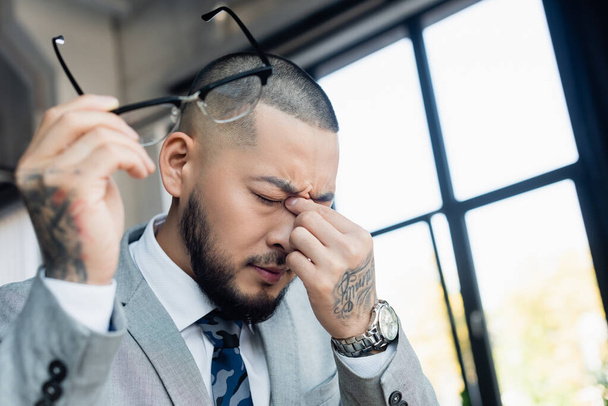 Erschöpfter asiatischer Geschäftsmann reibt sich die Augen, während er verschwommene Brillen im Büro hält - Foto, Bild