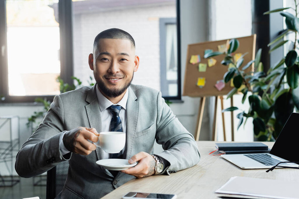 šťastný asijský podnikatel s šálkem kávy při pohledu na kameru, zatímco sedí na pracovišti - Fotografie, Obrázek