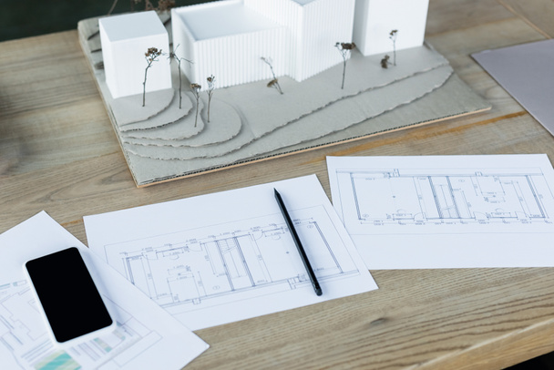 high angle view of architectural plans, house models and mobile phone with blank screen on wooden desk - Photo, Image