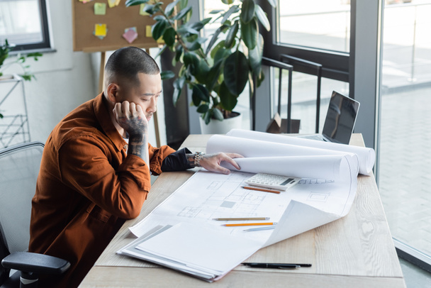задумливий азіатський архітектор, дивлячись на план під час роботи в будівельному бюро
 - Фото, зображення