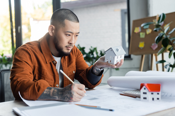 asian architect holding house model while drawing blueprint in office - Photo, Image