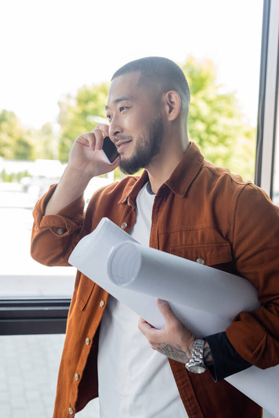 barbuto asiatico architetto con arrotolato cianografie sorridente durante telefono conversazione in ufficio - Foto, immagini