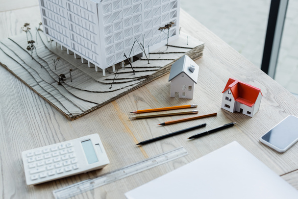house models, pencils, smartphone with blank screen and blurred calculator on wooden desk in office - Foto, afbeelding
