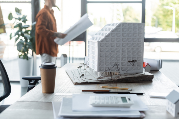 selective focus of house model and paper cup on desk near architect standing with paper on blurred background - Fotoğraf, Görsel