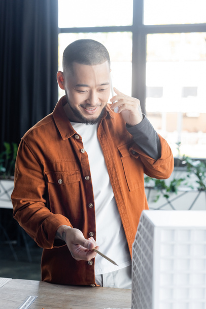 sorridente asiático arquiteto falando no smartphone e apontando com caneta em casa borrada modelo  - Foto, Imagem