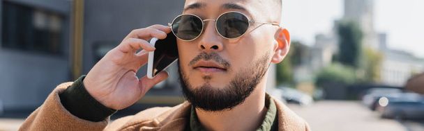 asian man in stylish sunglasses talking on smartphone outdoors, banner - Фото, изображение