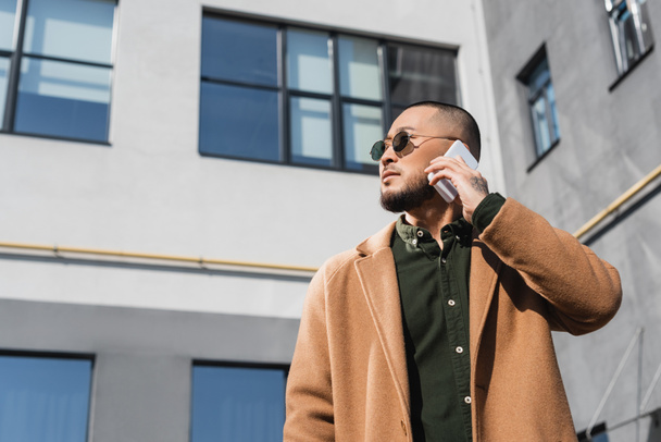 baixo ângulo vista de asiático homem em óculos de sol e casaco chamando no smartphone na cidade - Foto, Imagem