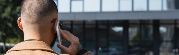 vue arrière d'un homme tatoué parlant sur un téléphone portable près d'un bâtiment flou, bannière - Photo, image