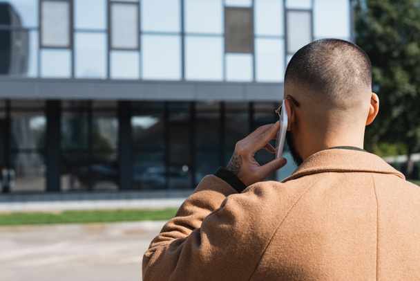 Rückansicht eines Mannes im beigen Mantel, der auf der Straße in der Stadt mit dem Handy telefoniert - Foto, Bild