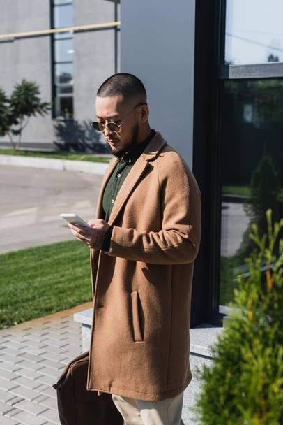 asian man in beige coat and sunglasses messaging on cellphone outdoors - Foto, imagen