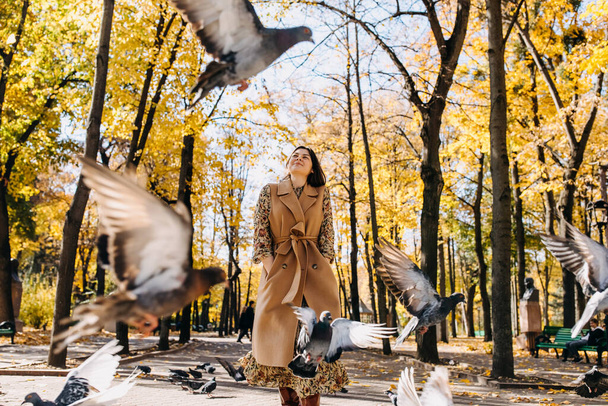 Młoda kobieta uśmiechnięta, spacerująca wśród gołębi w miejskim centralnym parku w ciepły jesienny dzień. - Zdjęcie, obraz