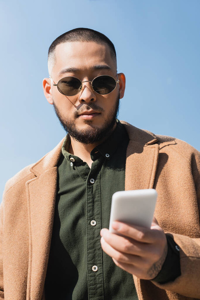 bajo ángulo vista de asiático hombre en abrigo y gafas de sol usando borrosa smartphone contra azul cielo - Foto, Imagen